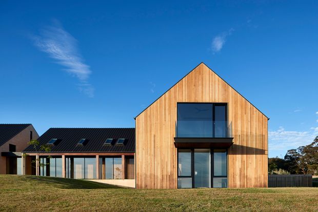 Exterior view of a contemporary house with a prominent steep gabled volume clad in warm vertical timber boards. Large dark-framed glazed openings include a recessed upper-level window and full-height lower doors, while a lower adjoining wing has a dark pitched roof and extensive glazing. The building sits in an open grassy setting under a clear blue sky.