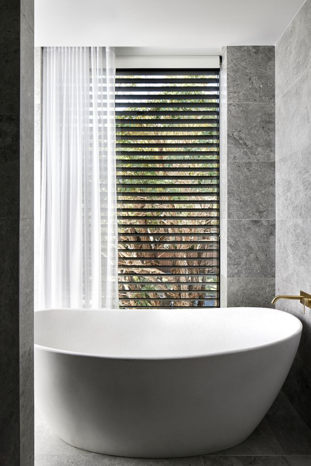 A refined contemporary bathroom featuring a large freestanding white oval bathtub against grey stone-look wall and floor tiles. A tall window with horizontal dark louvres and sheer white curtains brings in filtered natural light and views of greenery. A brushed brass wall-mounted tap provides a warm metallic accent.