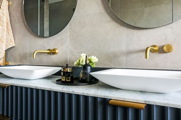 A refined contemporary bathroom vanity featuring two white vessel basins on a pale stone countertop, brushed brass wall-mounted taps, large rounded mirrors, and dark vertically fluted cabinetry with brass handles. Neutral grey wall tiles and sculptural basin forms create a luxurious, polished look.