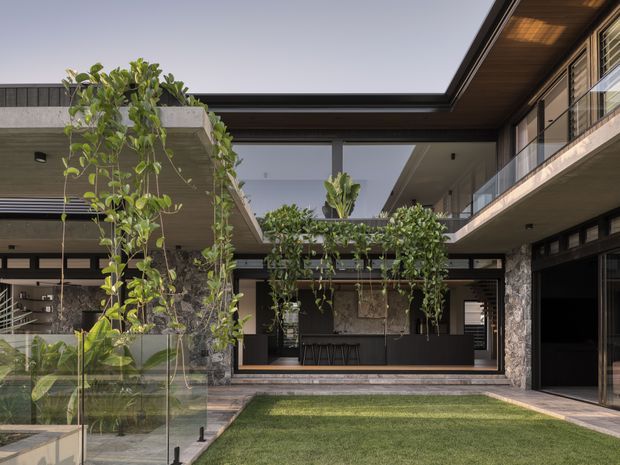 A sophisticated modern courtyard viewed from within a multi-level residence. The open-air central garden is framed by flat-roofed wings with extensive glazing, dark metal framing, stone-clad walls, timber soffits, and cantilevered upper-level balconies. A manicured lawn, glass balustrade, climbing greenery, and a covered outdoor entertaining area create an indoor–outdoor connection.