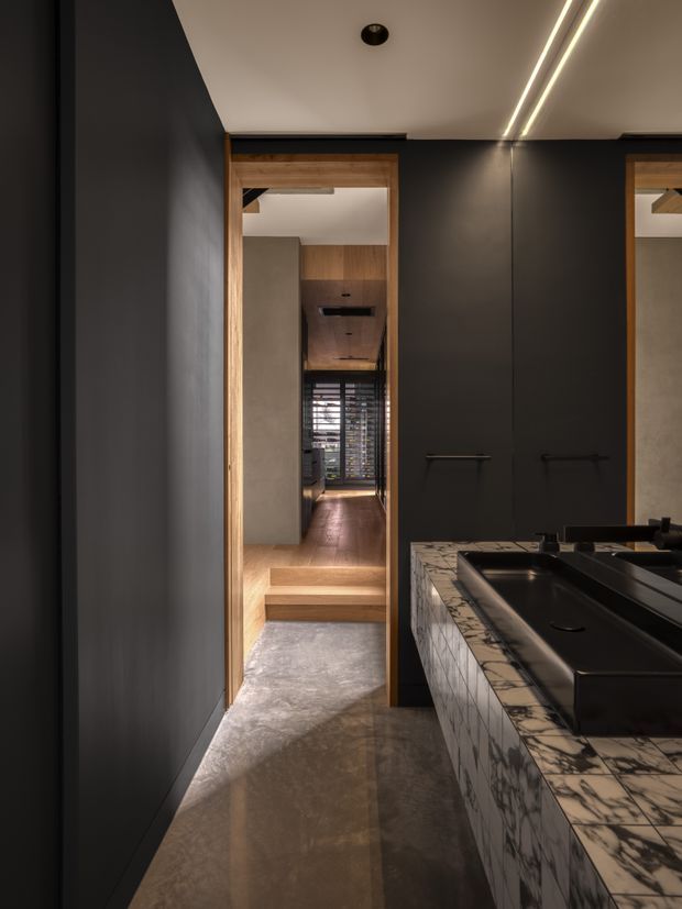 A narrow, dark-toned bathroom with a long black vanity and integrated basins on the right. The vanity is clad in a dramatic veined stone, while timber-framed doorways lead toward a raised hallway or adjoining area with timber flooring and shutters. Linear recessed lighting runs across the light ceiling, creating a refined contemporary contrast with the charcoal walls and polished concrete-like floor.