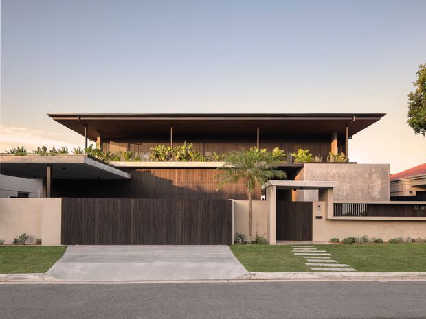 Street-facing view of a refined contemporary two-storey residence with a broad low-pitched roof, deep overhangs, warm timber screening, rendered and concrete-toned walls, and a strongly horizontal facade. The property includes a wide driveway, dark timber garage or entry gates, landscaped planting, a small front path, and a neatly maintained lawn.