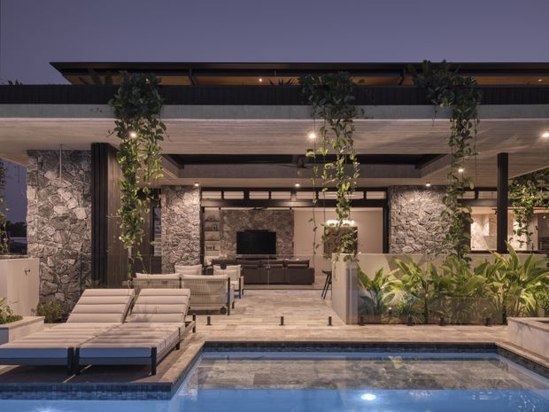 Twilight view of a sophisticated outdoor pool terrace adjoining an open-plan covered living area. The space features a rectangular swimming pool, light upholstered sun loungers, a large covered lounge with built-in or integrated seating and television, stone-clad walls, dark structural posts, timber-toned soffits, hanging greenery, and planted garden beds. Warm integrated lighting creates a calm, resort-like atmosphere.