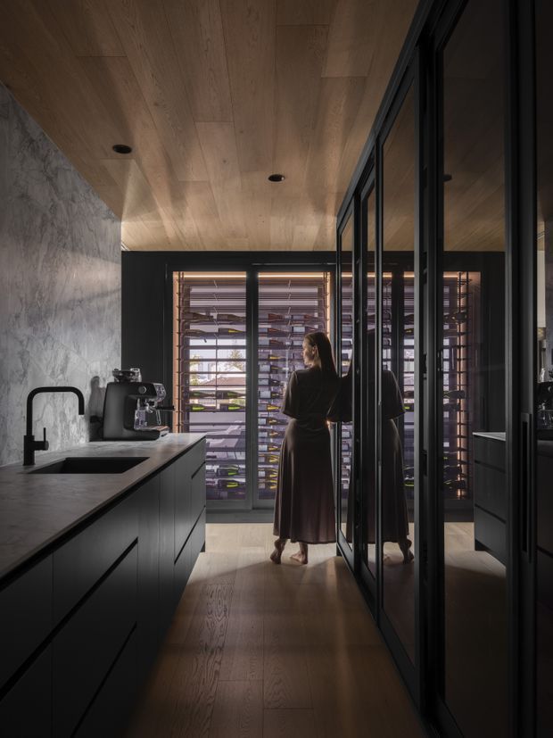 A moody, sophisticated kitchen interior viewed along a dark island toward a glazed wine storage room. The space features matte-black cabinetry, a dark stone benchtop and integrated sink, black-framed glass partitions, warm timber-lined ceiling and timber flooring, and a dramatic textured grey stone wall. A woman stands near the illuminated wine display, with reflections visible in the glazing.