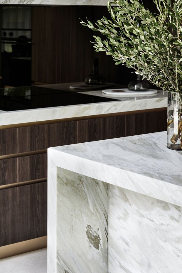Close-up view of a refined contemporary kitchen featuring dark timber cabinetry, a black induction cooktop, and prominent pale veined natural-stone surfaces on the island and rear bench. A large waterfall-edged island dominates the foreground, with sculptural dark vessels and leafy branches adding restrained decoration.