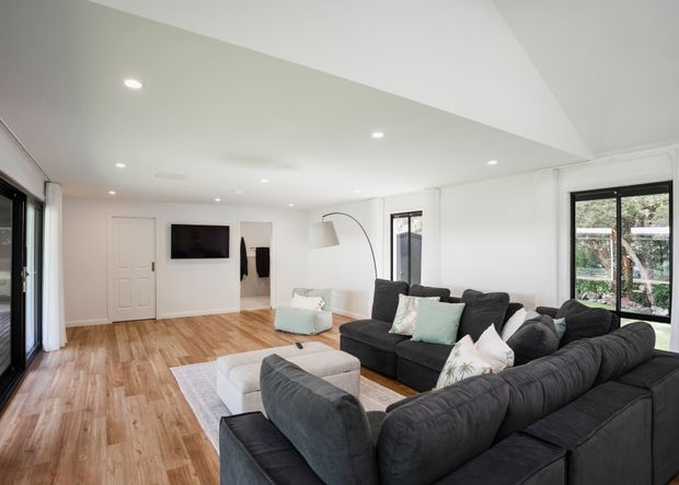 Bright, contemporary open-plan living room with white walls, recessed ceiling lighting, warm timber flooring, black-framed glazing, and a large dark sectional sofa arranged around a light rug and ottoman. A wall-mounted television, curved floor lamp, and doors to adjoining spaces are visible.