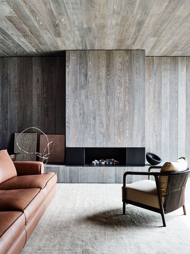 A refined living room with extensive vertically grained timber wall and ceiling panelling, a dark built-in fireplace surround, low linear joinery, a large pale rug, and mid-century-inspired leather and upholstered seating. The room has a warm, textural, contemporary character with strong contrast between light grey timber surfaces and darker timber accents.