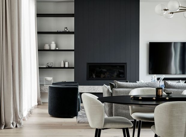 Refined open-plan living and dining area with a dark vertically panelled fireplace feature wall, built-in shelving, wall-mounted television, pale upholstered seating, a black oval dining table, and full-height sheer curtains. The space has a sophisticated neutral palette with layered textures and contemporary furnishings.