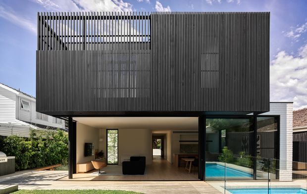 Exterior view of a contemporary two-storey residence with a dramatic dark vertical-screen upper facade projecting over a largely glazed ground floor. The open lower level connects to a covered outdoor living area and a visible swimming pool, while neighbouring light-coloured homes and a pitched roof are visible at the sides.