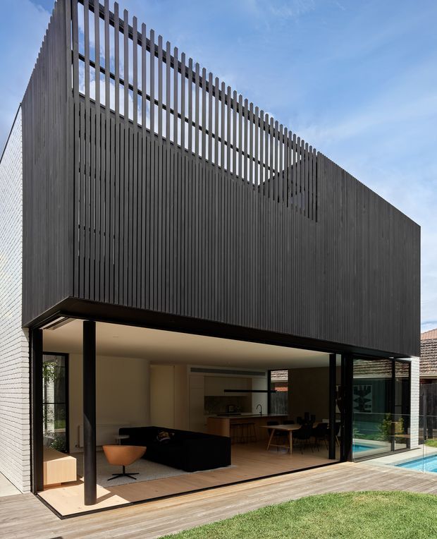 A contemporary two-storey residential extension with a dark vertically slatted upper facade and open ground-floor living area. Full-width glazing opens onto a timber deck and swimming pool, with a minimalist kitchen, dining area and living room visible inside. The projecting upper volume creates a sheltered outdoor zone, while the slatted screen continues above the roofline as a distinctive architectural feature.