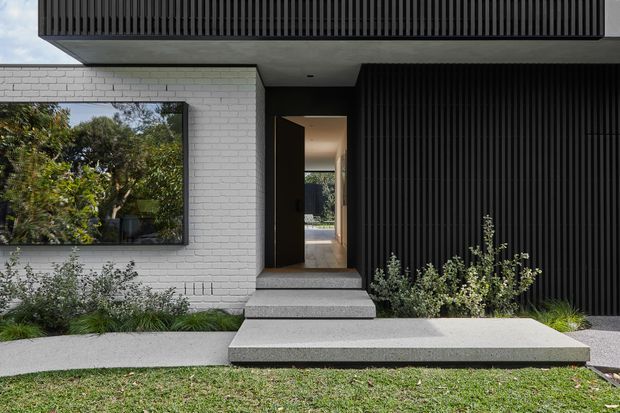 Contemporary residential front entrance with a white painted brick wall, dramatic black vertical-ribbed cladding, a projecting upper volume, and a recessed timber-toned doorway. Broad concrete entry steps and a minimalist landscaped garden create a clean, high-contrast facade.