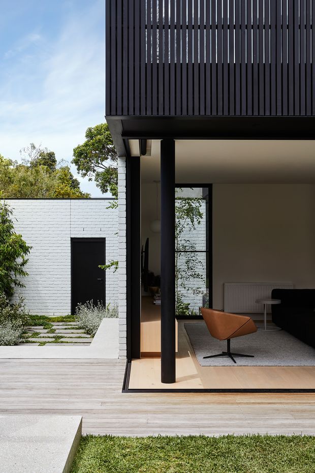 A contemporary house opening onto a timber deck and landscaped courtyard. The view highlights a dark vertical-slatted upper facade and balcony, slim black structural columns, expansive glazing, pale timber flooring, and a minimally furnished living area with a leather lounge chair, dark sofa, rug, and radiator. A white-painted brick courtyard wall and stepping-stone path are visible beyond.