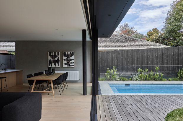A contemporary open-plan kitchen and dining area opening directly onto a timber pool deck through expansive black-framed glazing. The interior features pale timber flooring, dark cabinetry and ceiling elements, a timber dining table with black chairs, abstract wall art, and a minimalist linear kitchen. Outside, a rectangular swimming pool is bordered by grey paving, timber decking, a dark vertical-board fence, and established planting. The composition highlights the strong indoor-outdoor connection and clean modern detailing.
