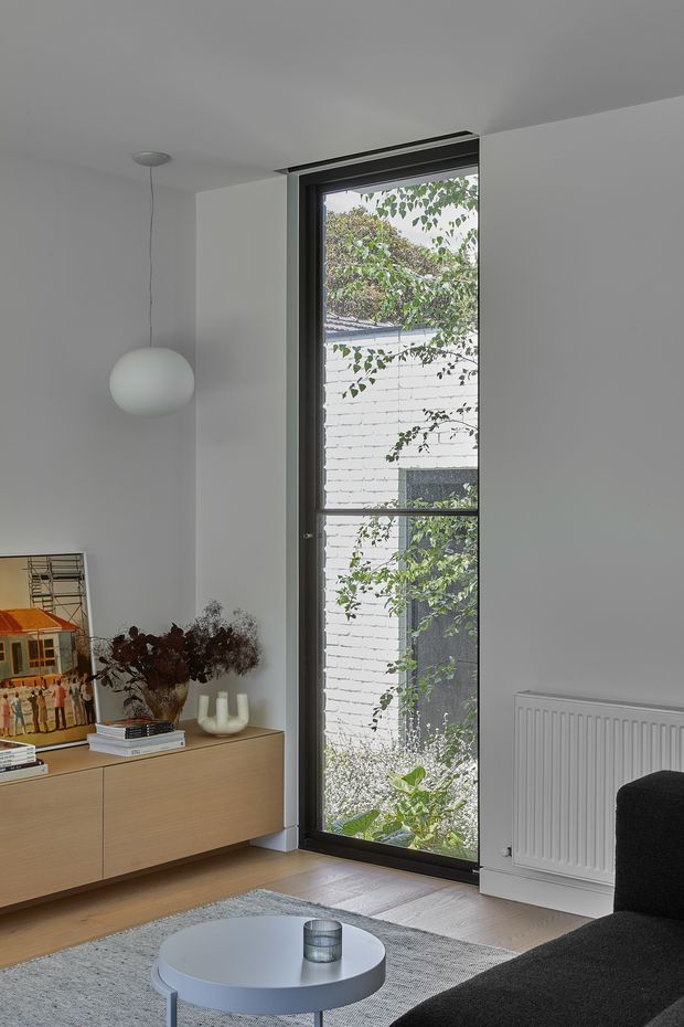 A compact, minimalist living room with white walls and ceiling, pale timber built-in cabinetry, and a tall black-framed glazed door or window opening to a leafy courtyard. The room includes a low timber sideboard with artwork and decorative objects, a black sofa, a round pale grey coffee table, a textured grey rug, a spherical pendant light, and a white wall radiator. The space is softly lit and restrained, with a strong connection to greenery outside.