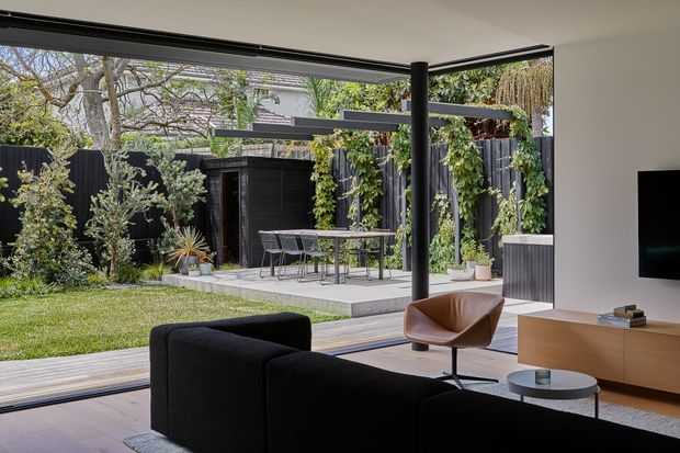 Contemporary living room opening onto a landscaped courtyard through wide floor-to-ceiling glazing. The space features a dark sectional sofa, leather lounge chair, low round coffee table, timber media console, and a covered outdoor dining terrace with climbing greenery and black fencing. The room has a clean, open connection between interior and garden, with slim dark window frames and a slender structural post.