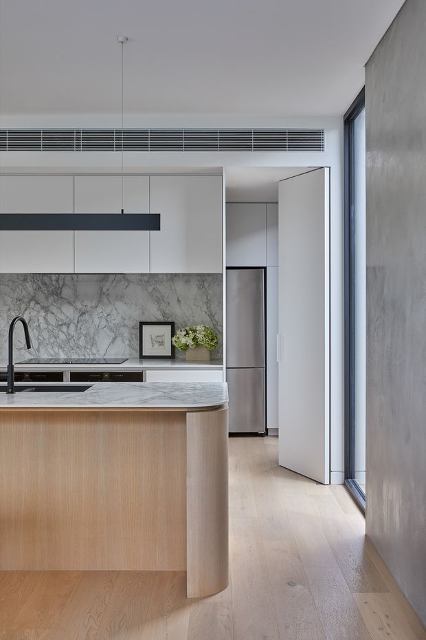 A refined contemporary kitchen viewed from the adjoining circulation area. The space features a large light-wood island with a curved end, a white-and-grey marble benchtop and full-height marble splashback, streamlined matte white cabinetry, integrated appliances, a black linear pendant light, pale timber flooring, and a tall glazed opening along the right side. A concealed or semi-open pantry/utility zone is visible beyond the kitchen.