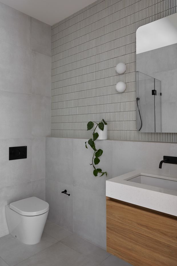 Contemporary bathroom featuring large-format grey wall and floor tiles, a narrow vertical grid-pattern tiled feature wall, a frameless glass shower, wall-hung toilet, black fixtures, and a floating timber-veneer vanity with a white integrated basin. A trailing plant and round wall lights add restrained natural detail.