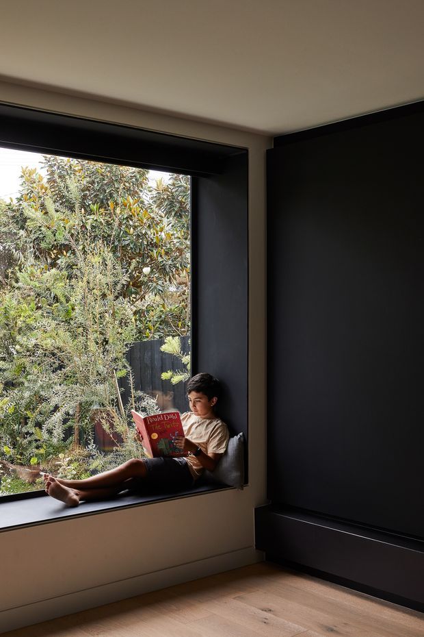 A quiet modern reading nook beside a large deep-set window, with a child seated on a built-in dark window seat reading. The room features a full-height matte-black built-in wall or cabinetry element, warm light timber flooring, neutral plaster walls, and abundant greenery visible outside. The composition is minimal, architectural, and softly lit.