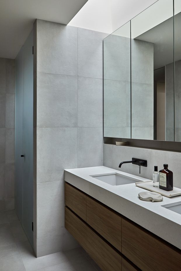A contemporary minimalist bathroom vanity with a long white integrated benchtop, twin rectangular basins, matte black wall-mounted tapware, and warm timber floating cabinetry. Large pale grey concrete-look wall tiles continue behind the vanity, while mirrored cabinets and a frameless glass shower enclosure create a clean, reflective finish. The room is softly lit with a concealed ceiling opening and has a restrained, refined palette.