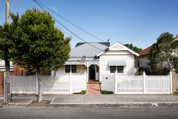 Street-facing view of a compact single-storey white weatherboard house with a pitched corrugated metal roof, decorative gabled porch detail, white picket fencing, and a narrow red-brick path leading to the entrance. Mature trees and neighbouring homes frame the residential facade.