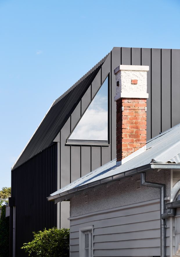 Exterior architectural detail showing a contemporary dark metal-clad extension rising behind an older weatherboard house. The extension has a steep gabled form, vertical standing-seam cladding, and a triangular window, contrasting with the original home's corrugated metal roof, painted weatherboards, exposed brick chimney, and rendered upper wall.