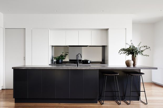 A bright, minimalist kitchen with a large dark island and integrated seating for two. White full-height cabinetry and a concealed appliance wall contrast with the dark island cabinetry and grey stone benchtop. Light timber flooring, simple recessed ceiling lights, a black tap and understated greenery create a clean contemporary interior.