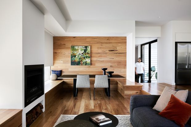 Bright contemporary open-plan living and dining area with a warm timber feature wall, integrated built-in dining nook, dark fireplace, timber flooring, and a charcoal sofa with layered cushions. Large glazing and a glass door bring in natural light, while clean white walls and a sculptural ceiling detail create a refined modern interior.