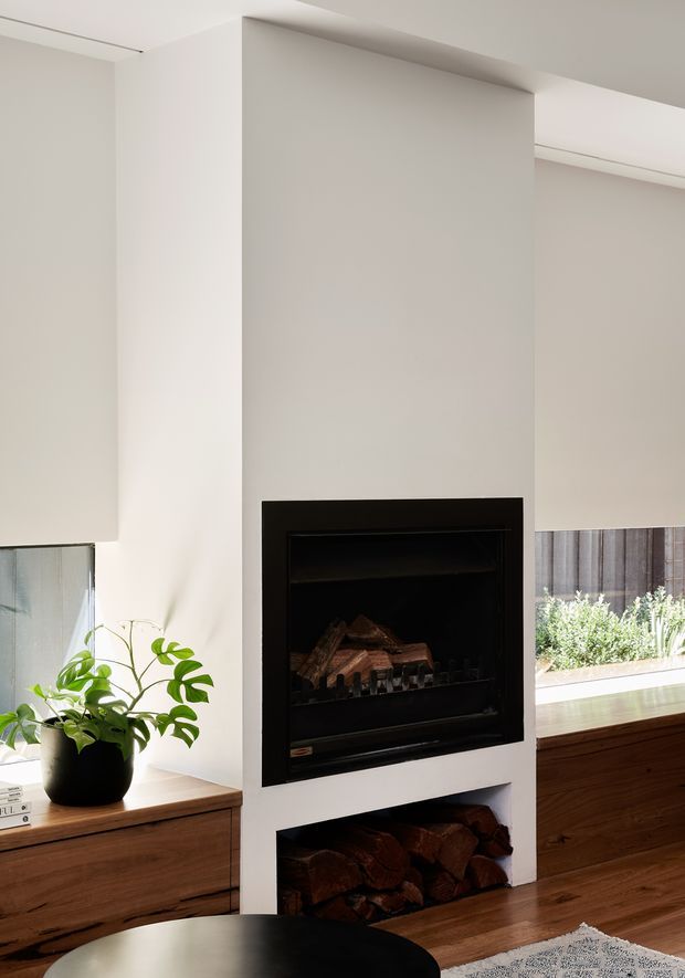 A bright, minimalist living room interior centred on a built-in black fireplace set into a white plaster feature wall, with an open timber log storage niche below. Low dark timber cabinetry runs along both sides, and a potted green plant sits near a window with a roller blind. The space has clean contemporary detailing and warm natural wood accents.
