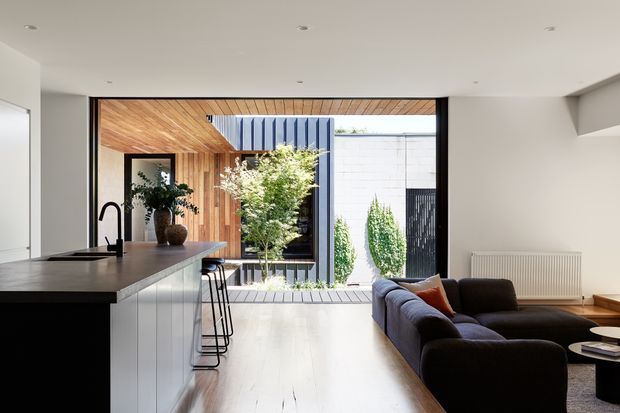A bright, contemporary open-plan living area combining a kitchen and lounge. The kitchen has a long dark benchtop with integrated sink, pale fixed cabinetry, and slim black bar stools. A dark sectional sofa occupies the living zone, with a small round coffee table and a radiator along the wall. Full-height glazing opens to a landscaped courtyard, while warm timber lining extends across the ceiling and a feature wall around the courtyard opening. The space has a clean, refined indoor-outdoor connection and abundant natural light.