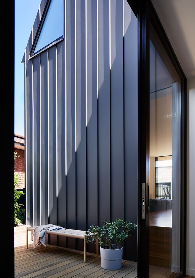 A close architectural view of a contemporary dark-clad building facade opening onto a timber deck or courtyard. Tall vertical ribbed metal panels create strong shadow lines, with a large black-framed glazed door revealing part of a refined interior. A simple timber bench, potted plant, and folded textile occupy the foreground, while a neighbouring brick wall is visible beyond.