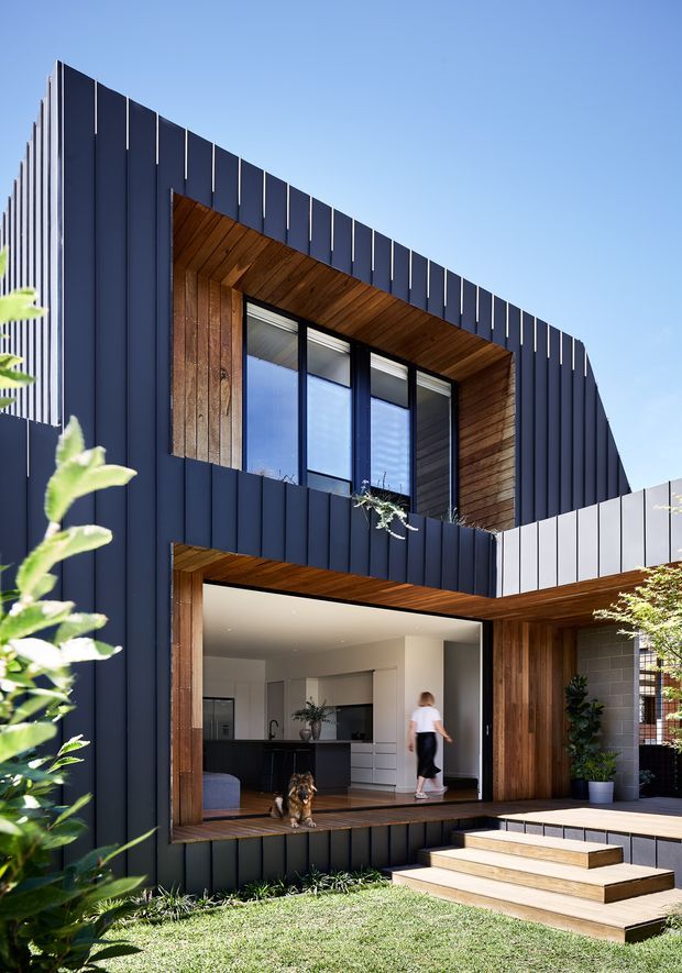 A contemporary two-storey residential facade with dark vertical metal cladding, warm timber-lined window reveals and soffits, and large black-framed glazing. The lower level opens onto a timber deck and stepped entry terrace, with a partially visible minimalist kitchen and living area inside. Landscaped planting and lawn frame the foreground.