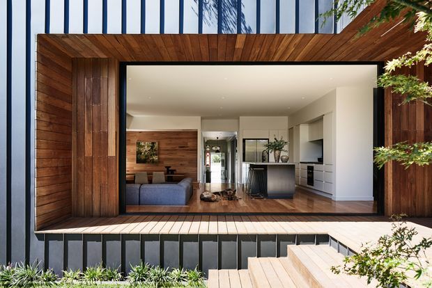 Contemporary residential home viewed from an exterior timber deck, looking through a large black-framed glazed opening into an open-plan living, dining and kitchen area. The facade combines warm timber cladding with dark vertical panels and extensive glazing. Inside are a blue-grey sofa, timber feature wall, kitchen island with dark base, white cabinetry and polished timber flooring. Timber decking, steps and planting frame the foreground.
