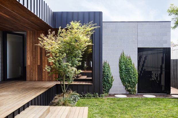 Contemporary residential exterior with a warm timber-clad entry and deck adjoining a dark metal-clad upper volume and a pale grey concrete-block wall. A tall black slatted pedestrian gate, vertical fencing, landscaped planting, and timber steps frame a compact lawn courtyard. The facade combines strong vertical screening with contrasting horizontal timber decking and masonry surfaces.