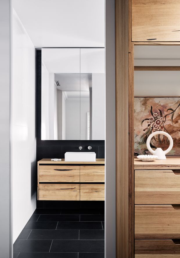 A narrow, contemporary bathroom viewed through a timber-framed opening. The bathroom features a floating natural timber vanity with four drawers, a white vessel basin, black wall tiles and black floor tiles, a large mirrored cabinet, and white walls. Adjacent built-in timber cabinetry with open shelving and artwork is visible at the right edge.