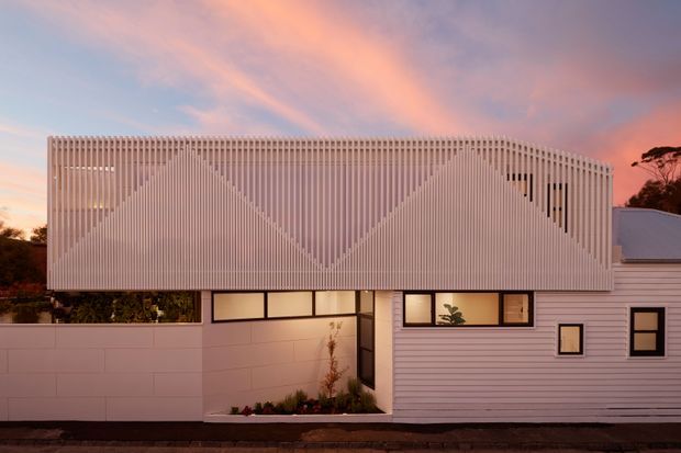 Exterior view of a contemporary multi-level residential building at sunset. The facade combines horizontal white cladding with a prominent upper-screen facade made of closely spaced vertical slats arranged in geometric triangular patterns. Dark-framed windows and glazed openings sit beneath the screen, with a small planted courtyard at ground level and a low-pitched roof visible to the right.