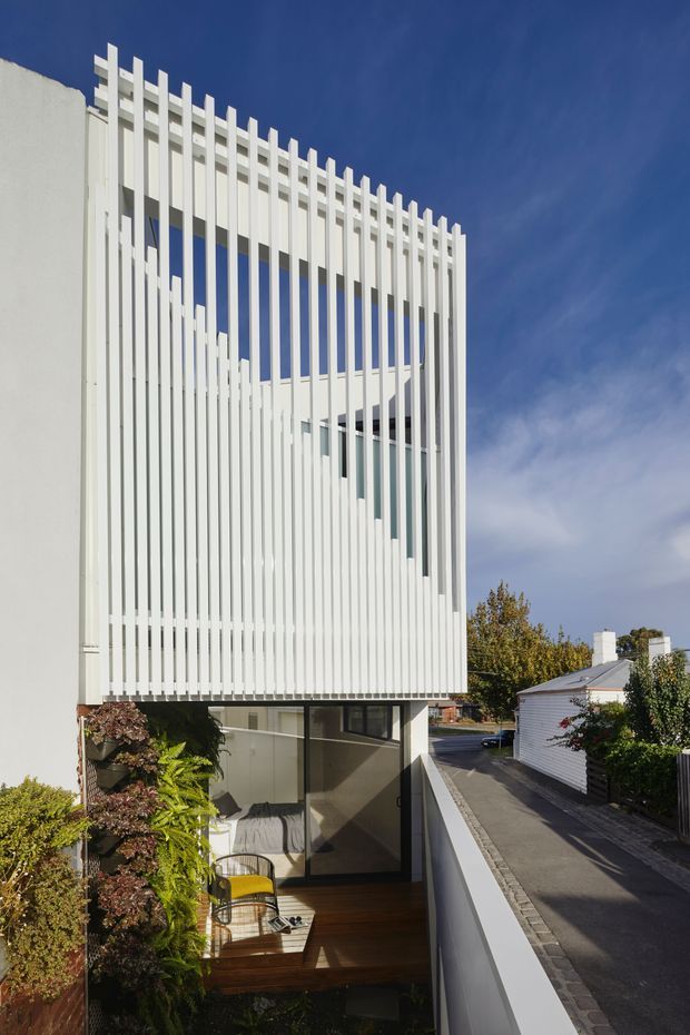 Exterior architectural photograph of a narrow, multi-storey contemporary building with a strong white vertical screen façade. Repetitive fins and projecting battens create a striking geometric pattern around the upper levels, with glazing and a recessed lower balcony or courtyard visible behind. The building sits along a narrow urban residential lane, with neighbouring low-rise structures and established planting.