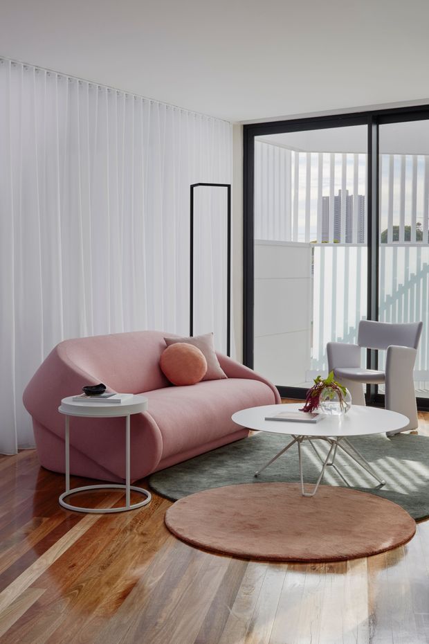 Bright contemporary living room with a curved blush-pink sofa, round white coffee and side tables, sculptural grey armchair, circular area rugs, and warm timber flooring. Full-height white vertical blinds and black-framed sliding glazing open to a balcony, with strong daylight and striped shadows adding a graphic effect.