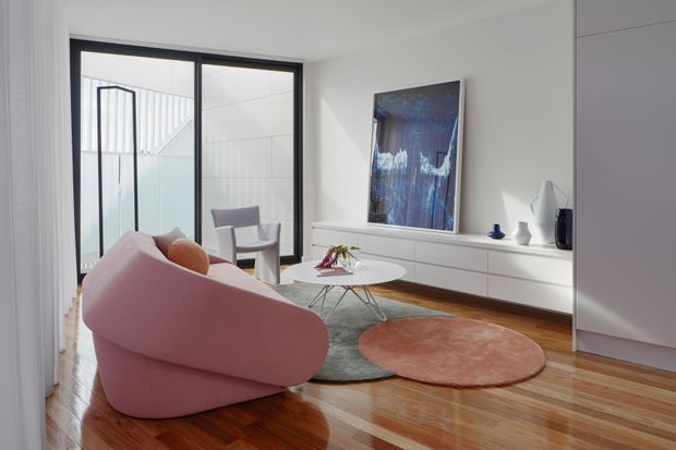 Bright contemporary living room with a sculptural blush-pink sofa, white low-profile built-in media cabinetry, round white coffee table, layered organic-shaped rugs, and polished timber flooring. Black-framed full-height glazing opens toward a stairwell, while a large abstract artwork and minimalist accessories add visual interest.