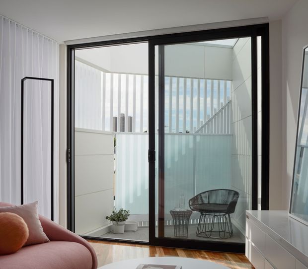 Bright contemporary living room opening onto a compact balcony or terrace through large black-framed sliding glass doors. The room includes a blush-pink sofa, white built-in cabinetry, timber flooring, and a small outdoor seating area enclosed by translucent privacy screening and vertical slats. Urban buildings are visible beyond the balcony.