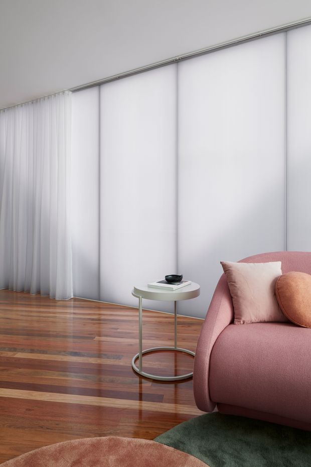 A softly lit contemporary living room corner with full-height translucent sliding or panelled window coverings, sheer white curtains, and polished warm timber flooring. A rounded blush-pink sofa with blush and terracotta cushions sits beside a small round side table, while a deep green rug and partial upholstered ottoman add colour and texture. The overall composition is refined, calm, and softly tonal.