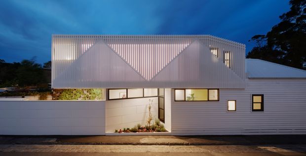 Twilight exterior view of a contemporary two-storey residence with a long, horizontally composed lower volume and a distinctive upper box wrapped in translucent vertical ribbed cladding. Warm interior lighting glows through narrow windows and the screened facade, with a sheltered entry courtyard, pale boundary wall, and planted strip visible at street level.