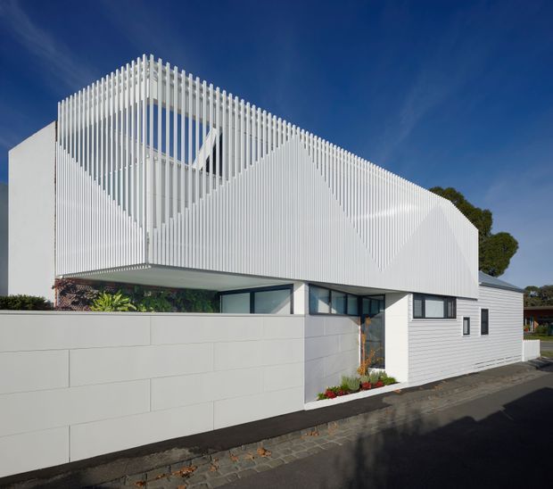 Street-level exterior view of a highly contemporary single-storey residence. The building has crisp white geometric roof and wall forms, extensive vertical white screening fins forming a sculptural upper facade, dark-framed glazing, white horizontal cladding, and a low pale masonry boundary wall. A paved footpath and small planted entry garden sit in front, with a clear blue sky and a glimpse of neighbouring roofs in the background.