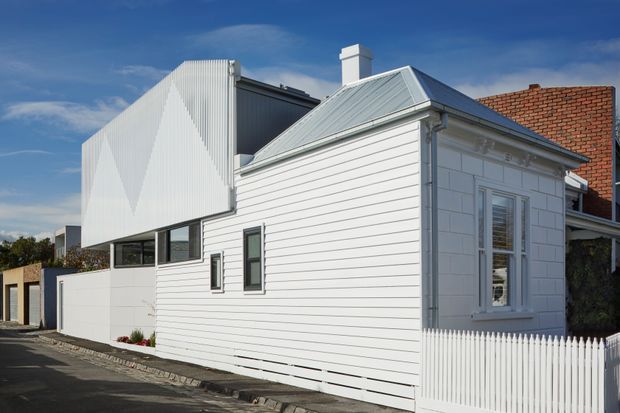 Street-facing exterior view of a renovated or extended residence combining a traditional white weatherboard cottage with a sharply contemporary rear addition. The cottage has a low-pitched metal roof, decorative window trim, chimney, and white picket fence, while the addition features bold faceted white cladding and dark-framed glazing. A neighbouring brick structure is partially visible.