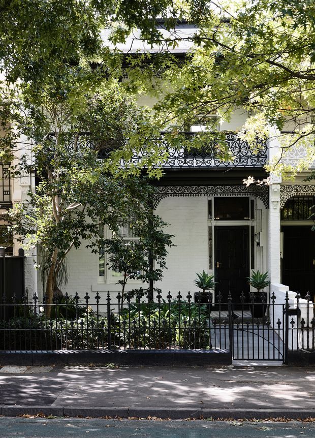 Street-facing view of a narrow, multi-storey terrace house with a white-painted masonry facade, black decorative wrought-iron balcony balustrades, ornate ironwork above the entry, black front door and gate, and a small landscaped front garden behind a low iron fence. Mature tree canopy partially frames the facade.