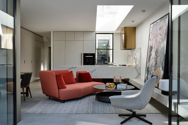 A bright, open-plan contemporary living room with a compact integrated kitchen at the rear. The space features a sculptural coral-red sofa, grey lounge chair, black oval coffee tables, pale cabinetry, a marble-look kitchen island and splashback, a large abstract artwork, a skylight, and extensive dark-framed glazing.