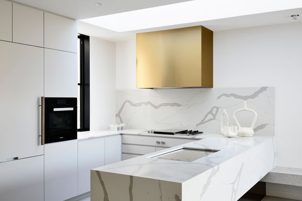 Bright contemporary kitchen with white flat-panel cabinetry, integrated black appliances, a large marble-look island, matching waterfall benchtop and splashback, and a prominent brushed gold rangehood enclosure. A linear skylight or ceiling opening adds natural light.