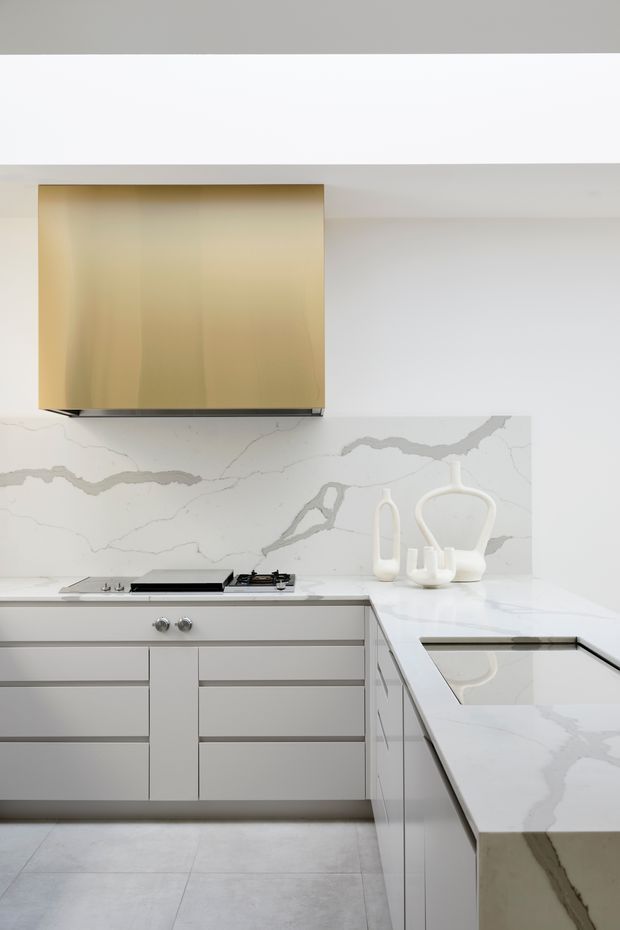 A bright, minimalist contemporary kitchen with a white and pale grey palette. The compact L-shaped layout features flat-panel light cabinetry, a white marble-look stone benchtop and matching full-height splashback with prominent grey veining. A brass-gold rangehood provides the main visual accent above a gas cooktop, while a pale tiled floor and integrated appliances maintain a clean, streamlined appearance.