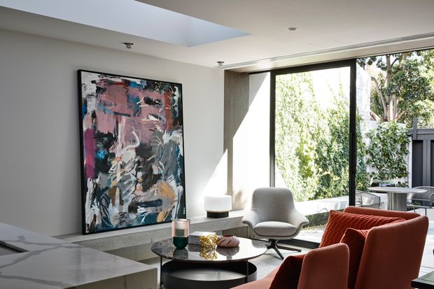A refined contemporary living room with a large abstract artwork, sculptural lounge chair, round black coffee tables, and rust-coloured upholstered armchairs. Floor-to-ceiling black-framed glazing opens the room to leafy greenery, while a broad skylight and pale walls create a bright, airy interior. A pale stone fireplace or built-in ledge is visible beneath the artwork.