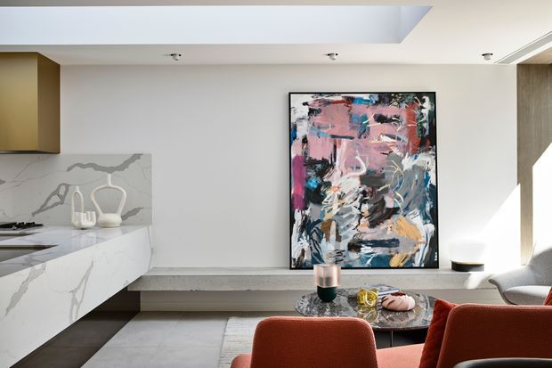 A bright, contemporary open-plan kitchen and living area. The kitchen features a white marble-look island and splashback, pale cabinetry, and integrated appliances. A built-in grey bench runs along the wall beneath a large colourful abstract artwork, with red upholstered dining or lounge chairs and a low coffee table in the foreground. Recessed ceiling lighting, streamlined joinery, and strong natural light create a refined, modern interior.