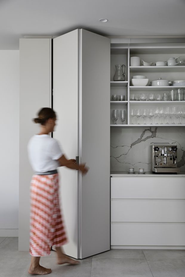 A minimalist white kitchen focused on a tall pivoting or folding pantry cabinet beside open shelving. The shelving displays white ceramics and glassware above a built-in espresso machine, with a veined white marble backsplash and handleless white drawers below. A person is shown opening the tall cabinet. The room has a pale grey tiled floor, smooth white cabinetry, and a restrained monochromatic aesthetic.
